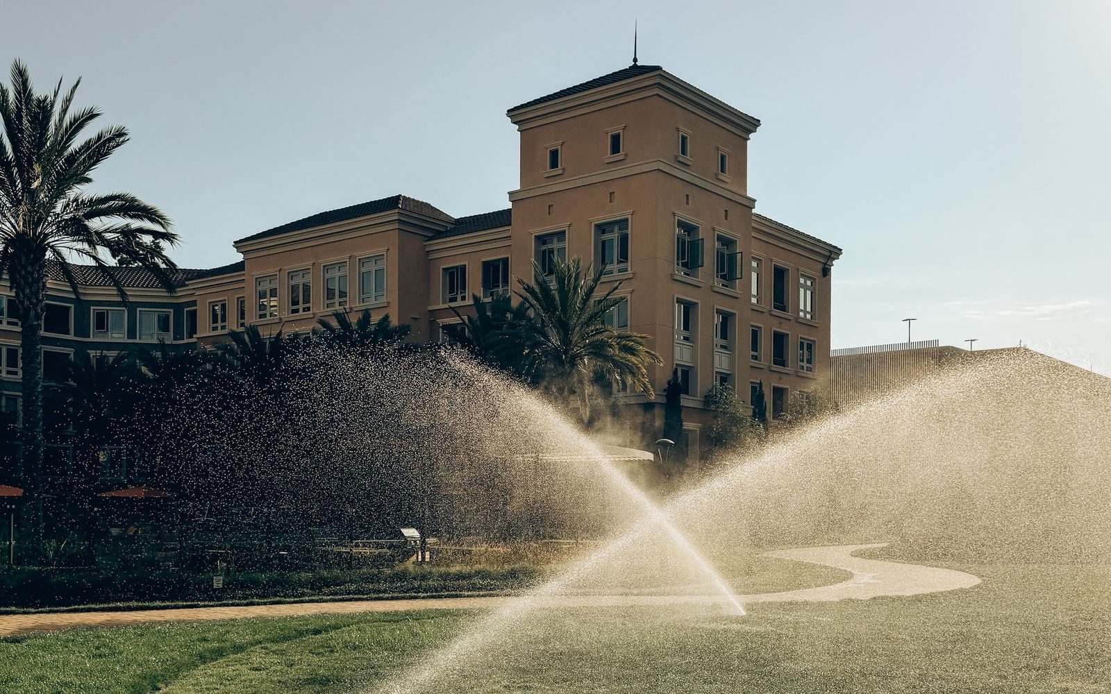 Rotors watering a lawn in front of a tile-roof building and palms.